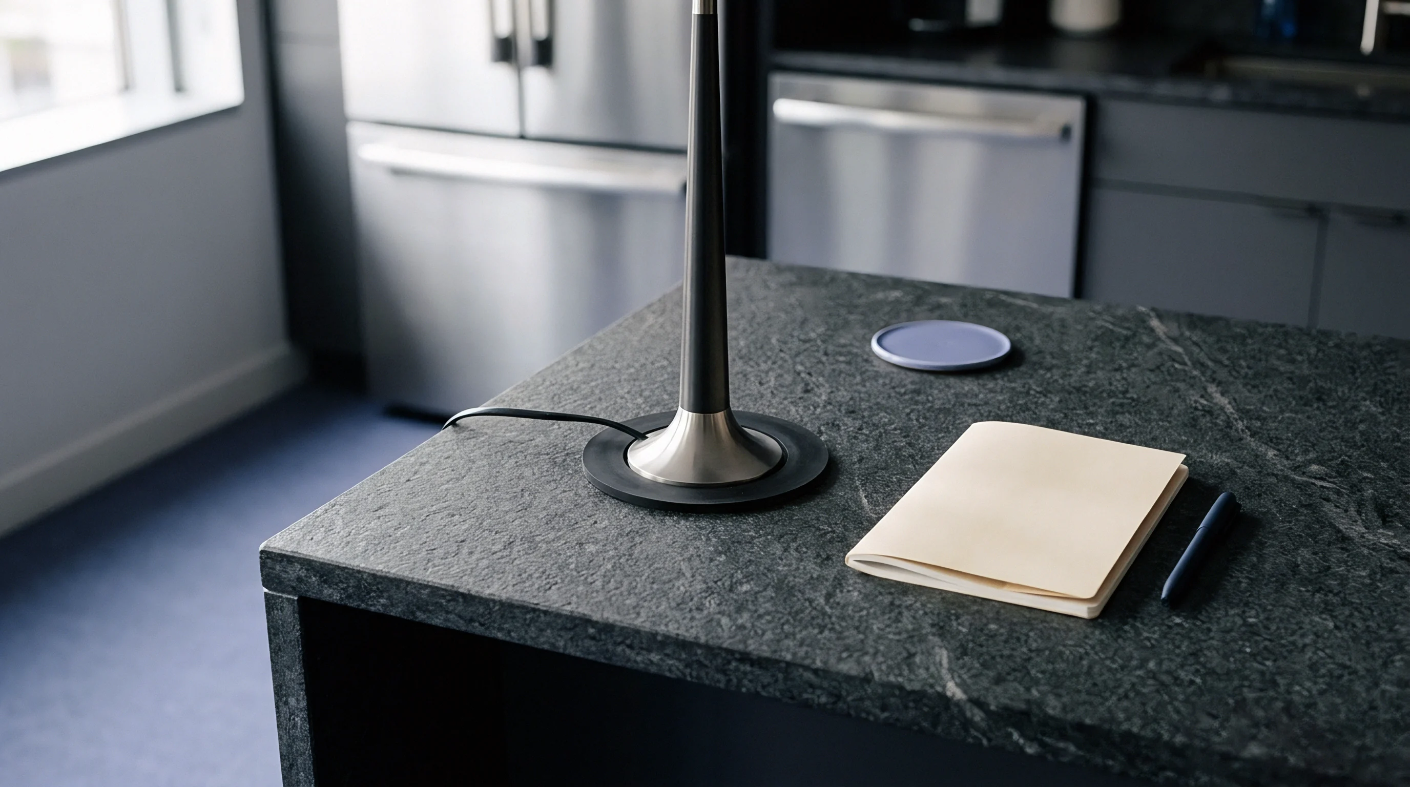 A dark metal task lamp stands beside a bound paper notebook and a blue pen upon a textured granite countertop surface.