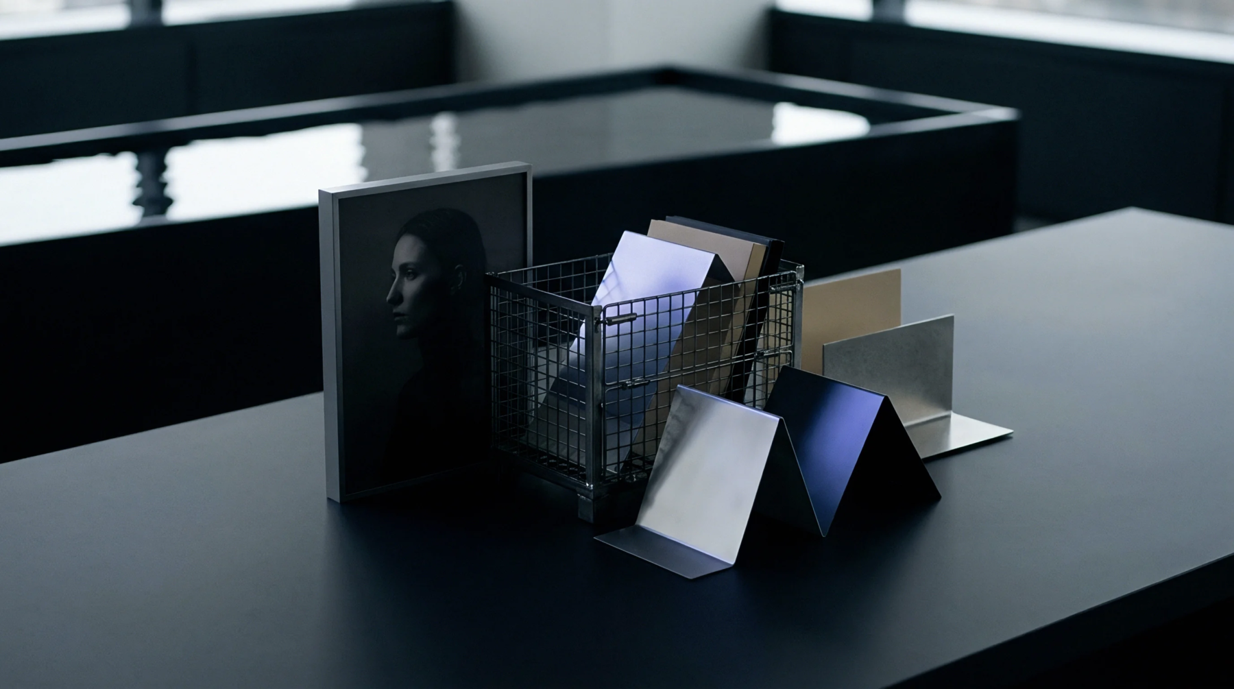 Metal finishes and polymer sheets rest inside a wire mesh bin beside a portrait print upon a dark assembly table surface.
