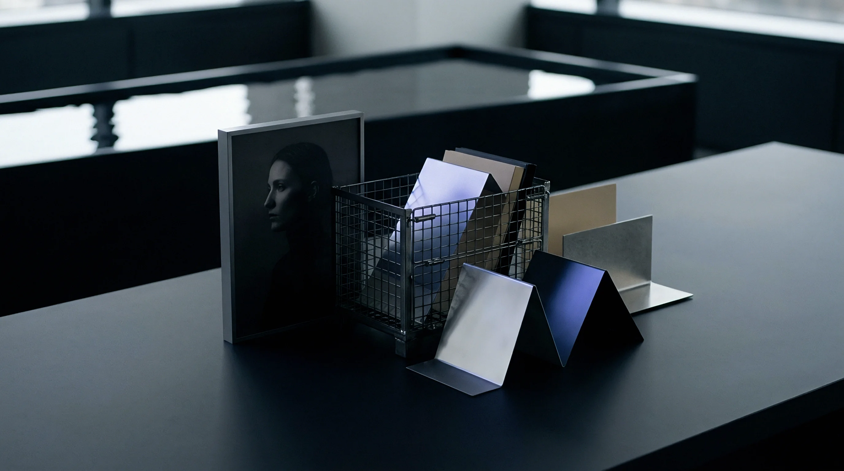 Metal finishes and polymer sheets rest inside a wire mesh bin beside a portrait print upon a dark assembly table surface.
