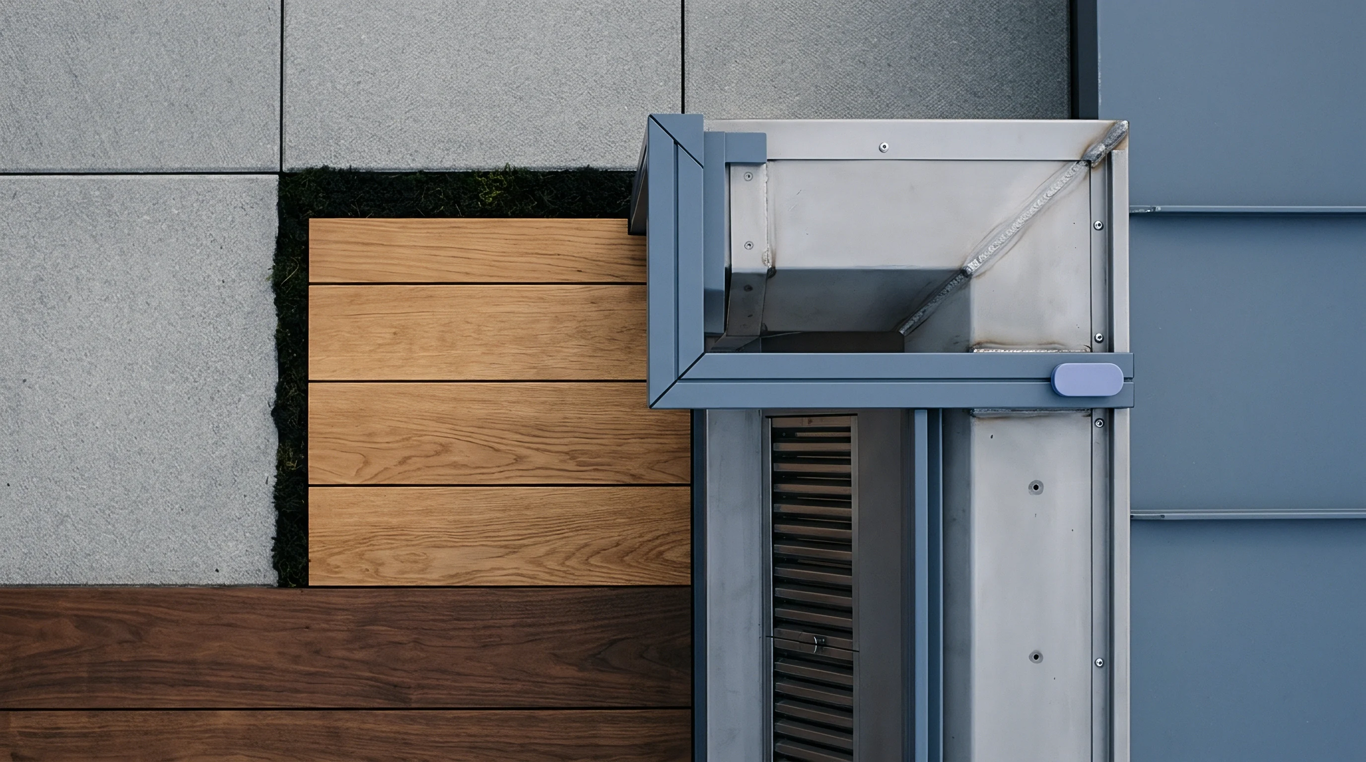 Grey stone tiles and wood planks abut a brushed metal structure with a blue metal frame integrated with a patch of dark ground cover.