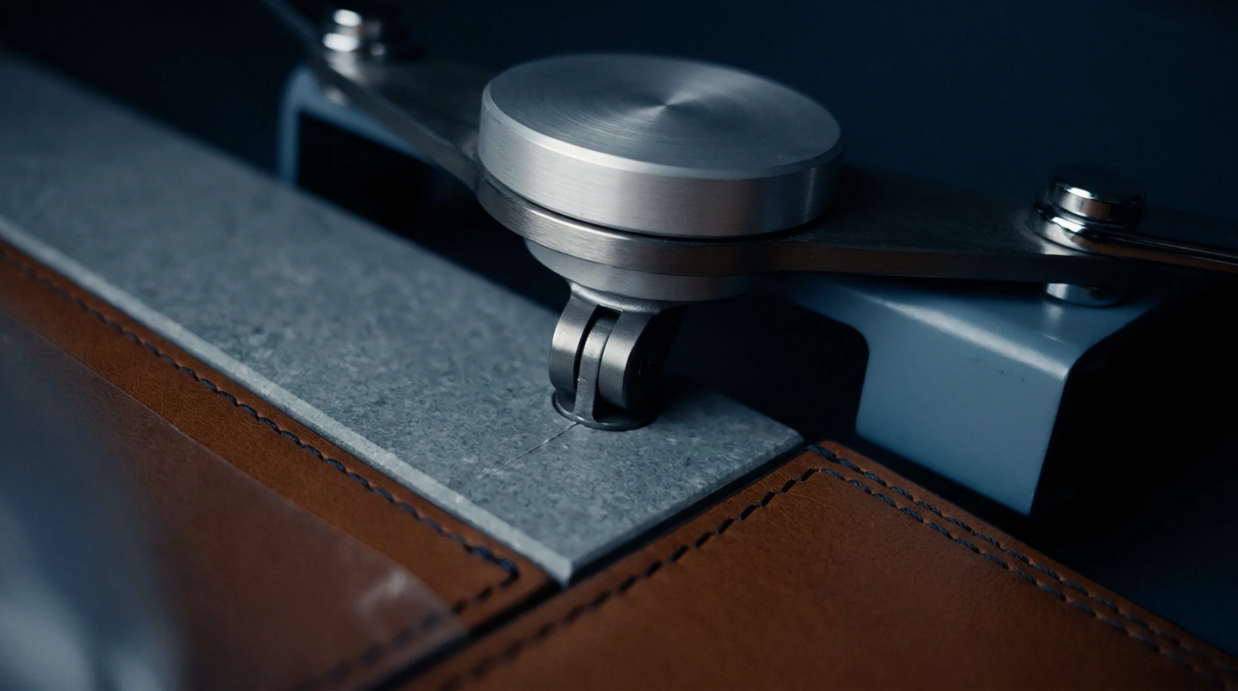 A turned stainless steel hinge mechanism secures a gray stone plate adjacent to stitched brown leather panels on an industrial assembly.